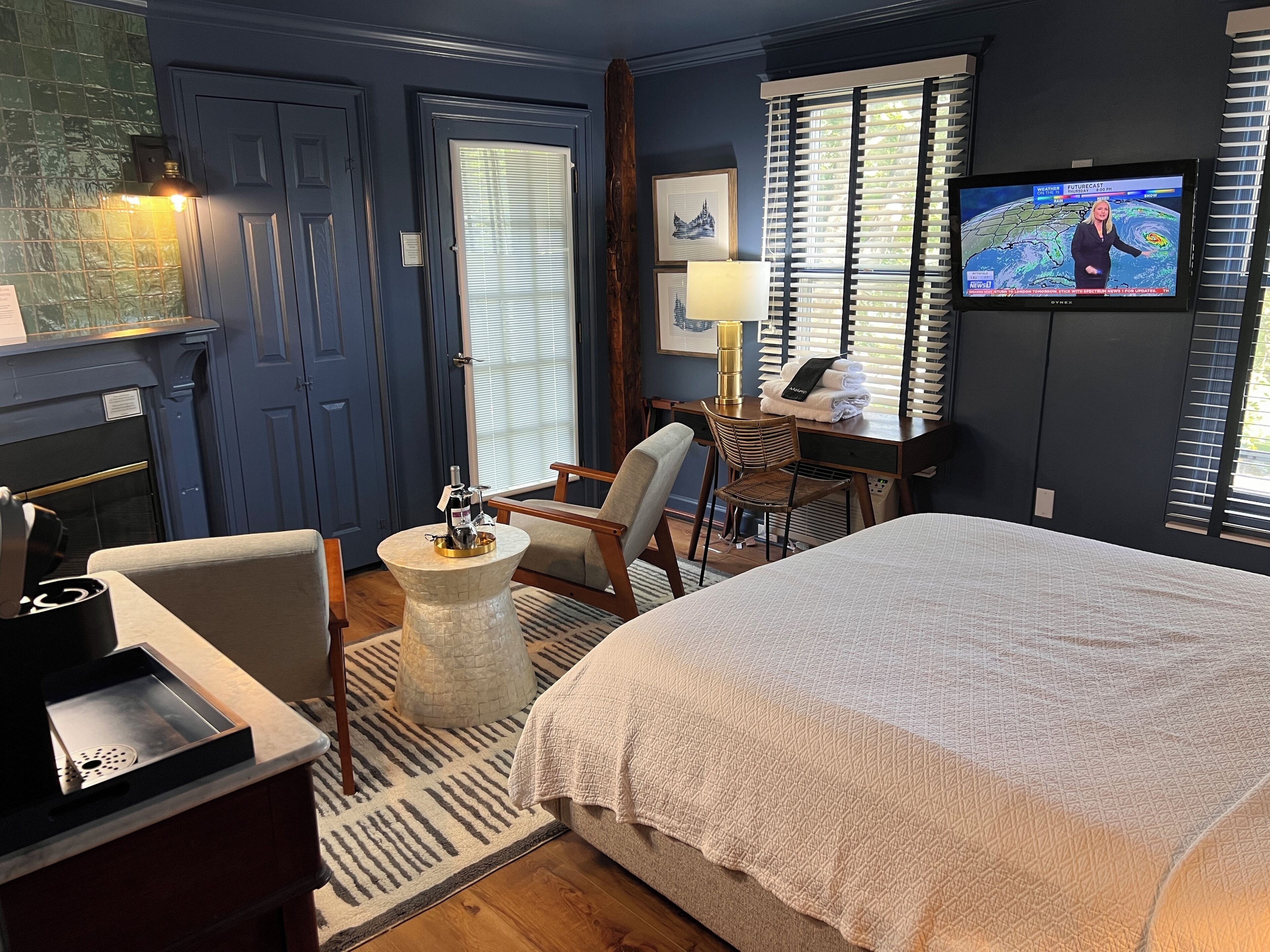 A hotel room with a bed, a desk with a chair, a TV, and a fireplace.