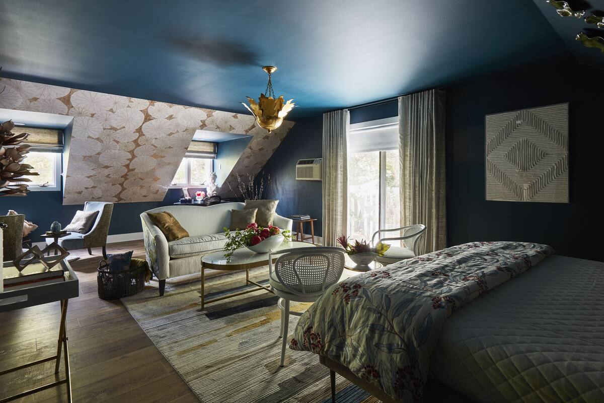 Un dormitorio moderno con una cama grande, un sofá y una mesa de comedor. Hay ventanas con cortinas, una araña y un adorno en la pared.
