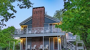 Uma casa de dois andares com varanda, chaminé de tijolos e um alpendre com cadeiras Adirondack.