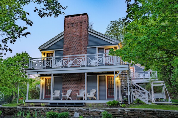 A two-story house with a balcony, a brick chimney, and a porch with Adirondack chairs.