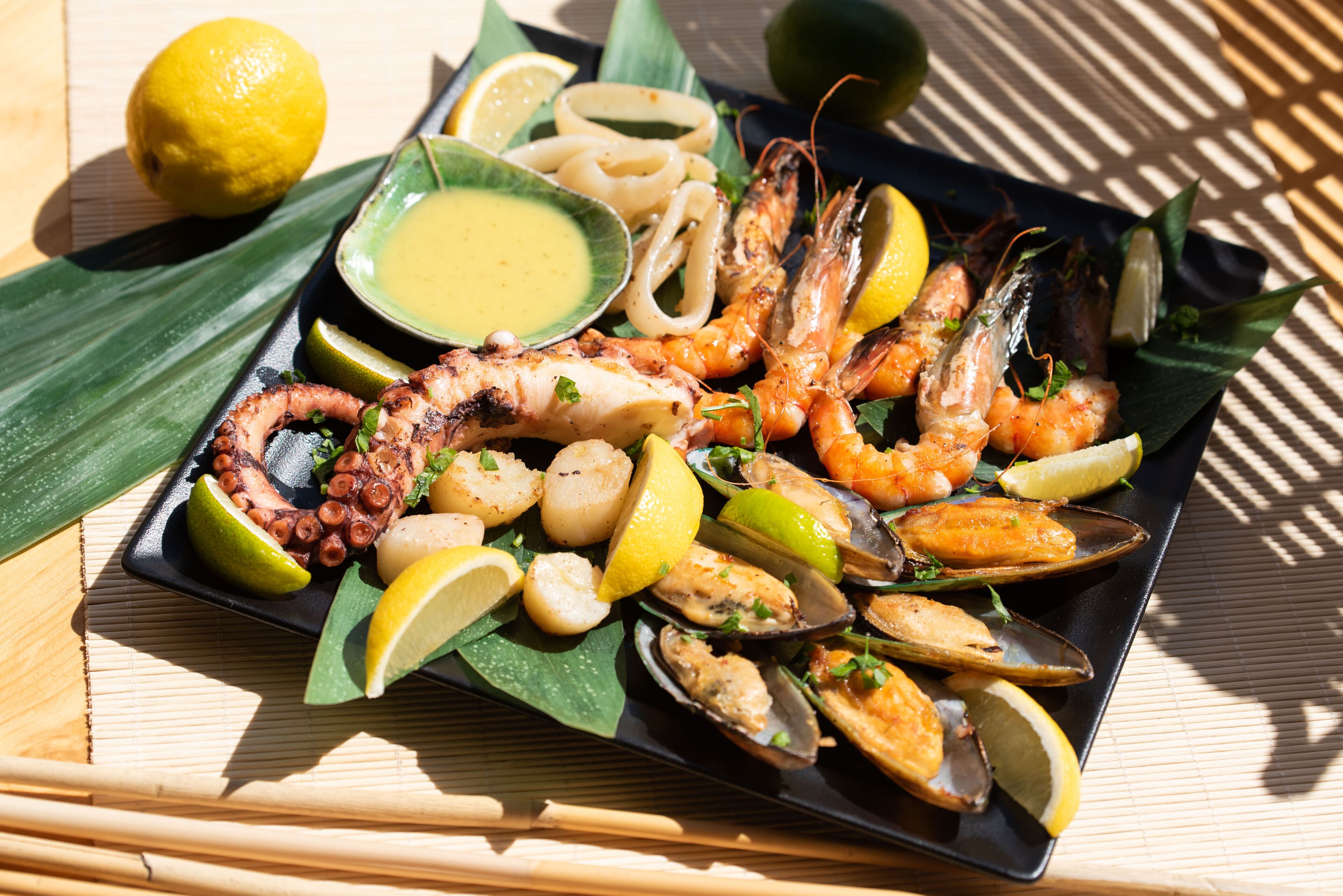 A seafood platter with shrimp, mussels, and squid, garnished with lemon slices and herbs, served on a black plate with a side of sauce.