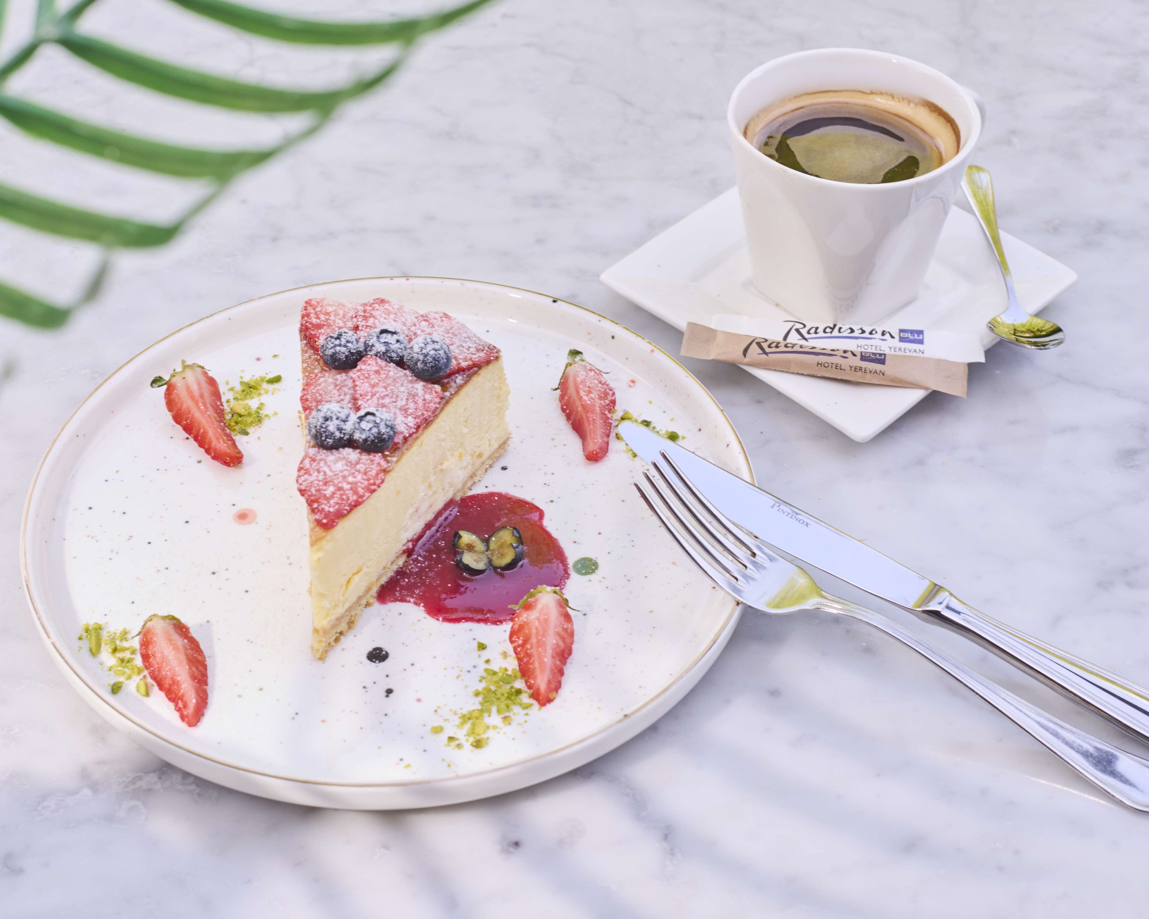 A slice of cheesecake with strawberries and blueberries on a plate, a cup of coffee, and a fork on a marble surface.