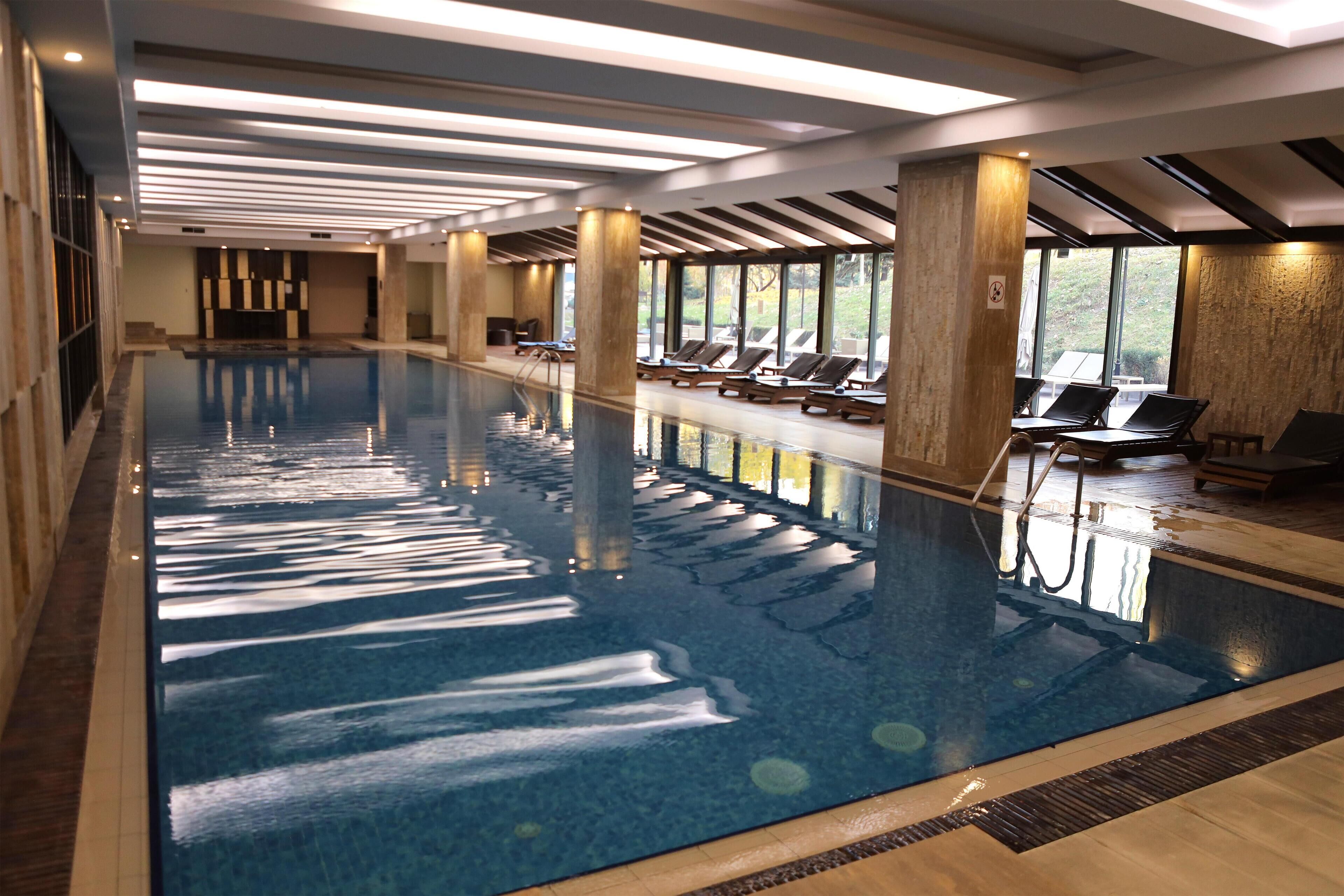 An indoor swimming pool with a long, narrow shape, surrounded by lounge chairs and large windows.
