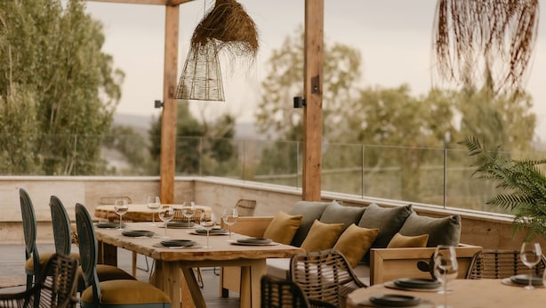 Un área de comedor al aire libre con mesas y sillas de madera, rodeada de vegetación y vistas a árboles.