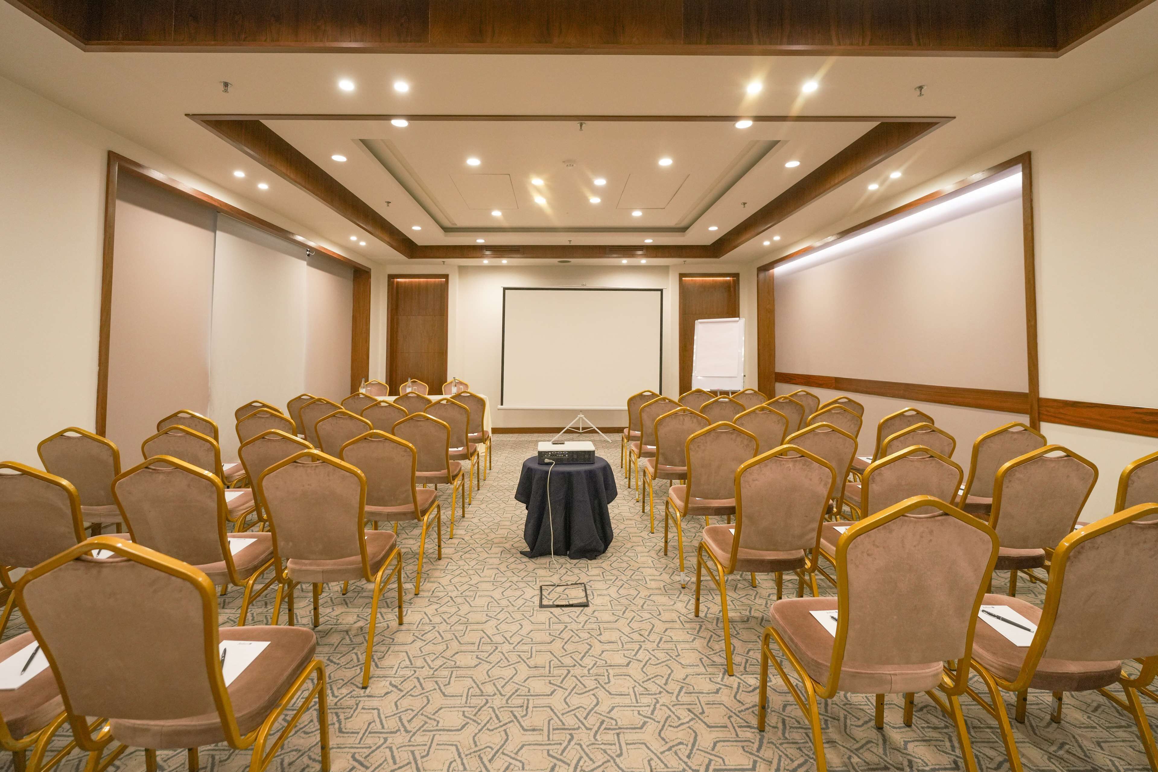 Un salón de conferencias con filas de sillas, una pantalla de proyector y una pizarra.