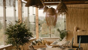 Un comedor con una mesa larga dispuesta para una comida, rodeada de sillas de mimbre, con ventanas grandes y vistas a la vegetación exterior.