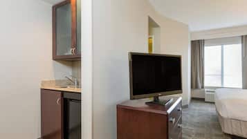 A hotel room with a flat-screen TV on a wooden dresser, a small kitchenette with a sink, and a large window with curtains.