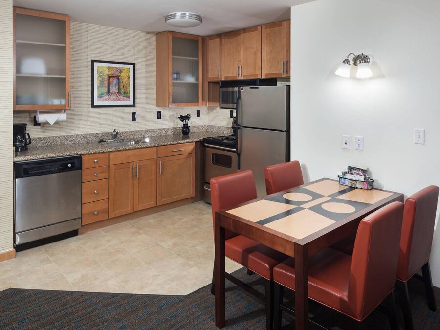 A compact kitchen with wooden cabinets, a stainless steel refrigerator, a microwave, and a dining table with red chairs.