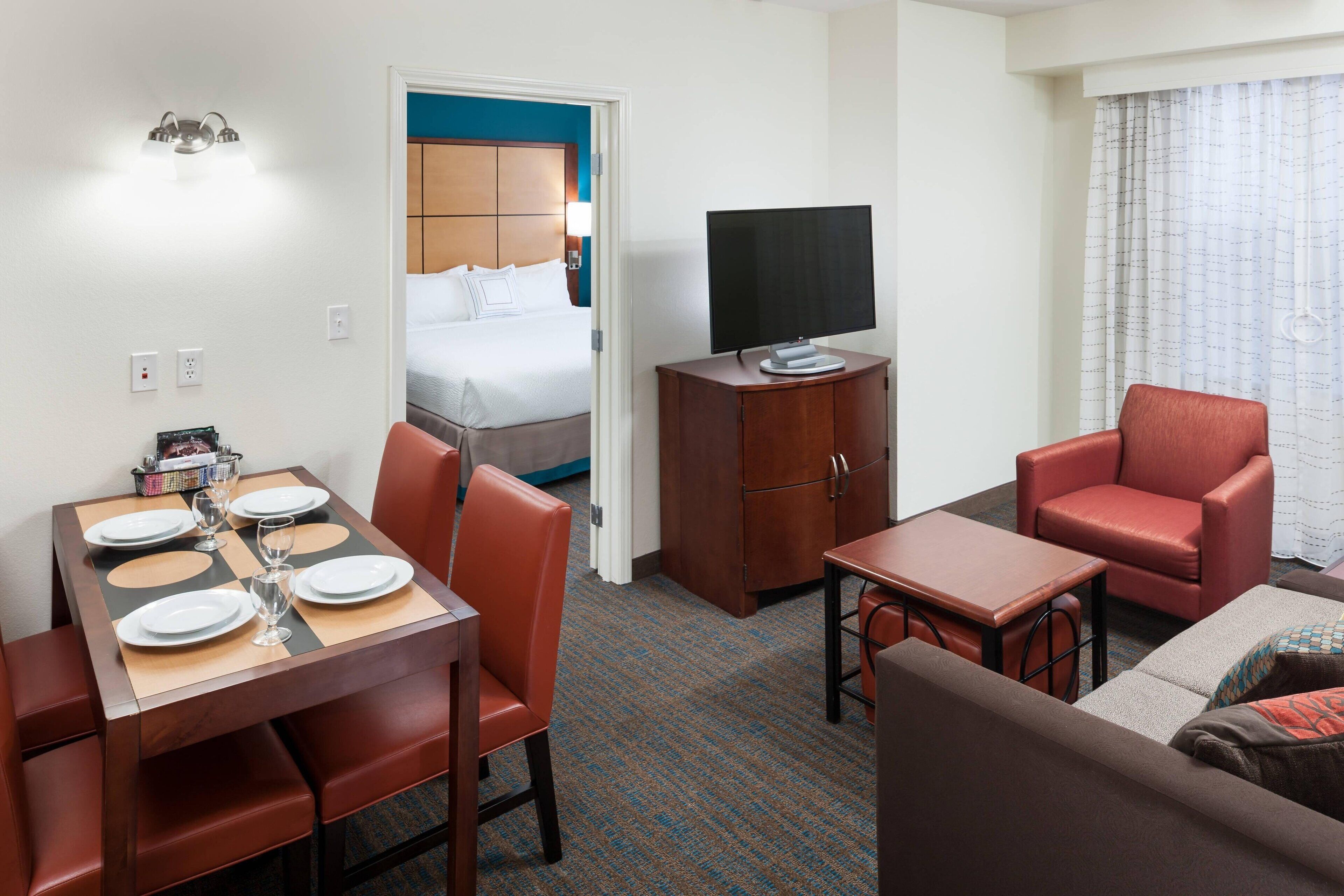A hotel room with a bed, a TV, a dining table with chairs, a sofa, and a small table with a lamp.