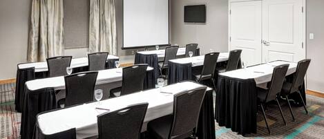 A conference room with a large screen, multiple tables with chairs, and a carpeted floor.
