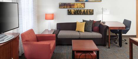 A modern hotel room with a flat-screen TV, a sofa, a red armchair, a small wooden table, and a desk with a chair.