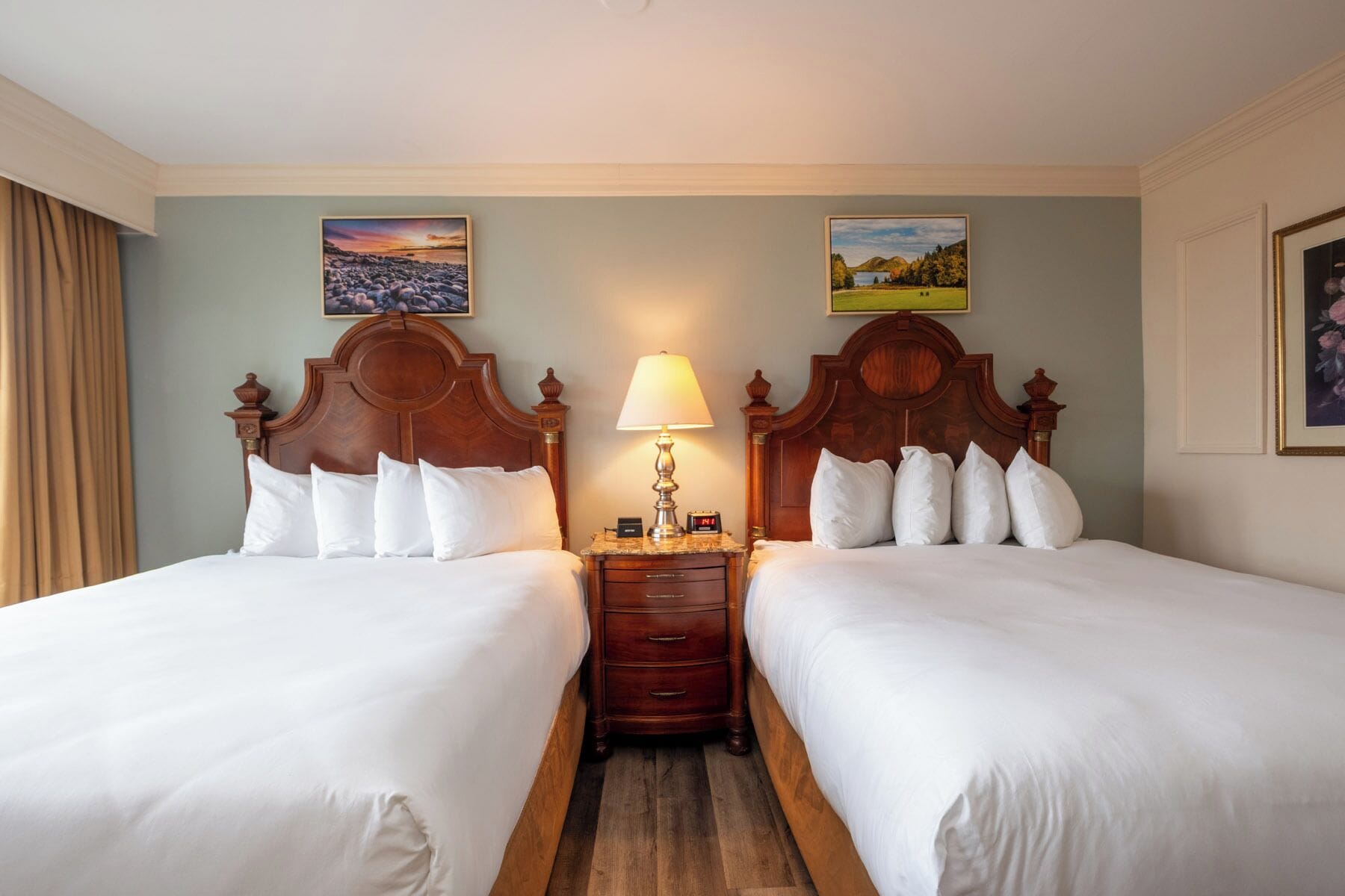 A hotel room with two beds, a nightstand with a lamp, and two paintings on the walls.