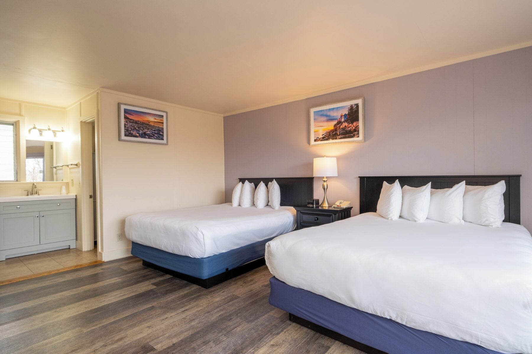 Quarto de hotel com duas camas, banheiro com espelho e duas quadros na parede.