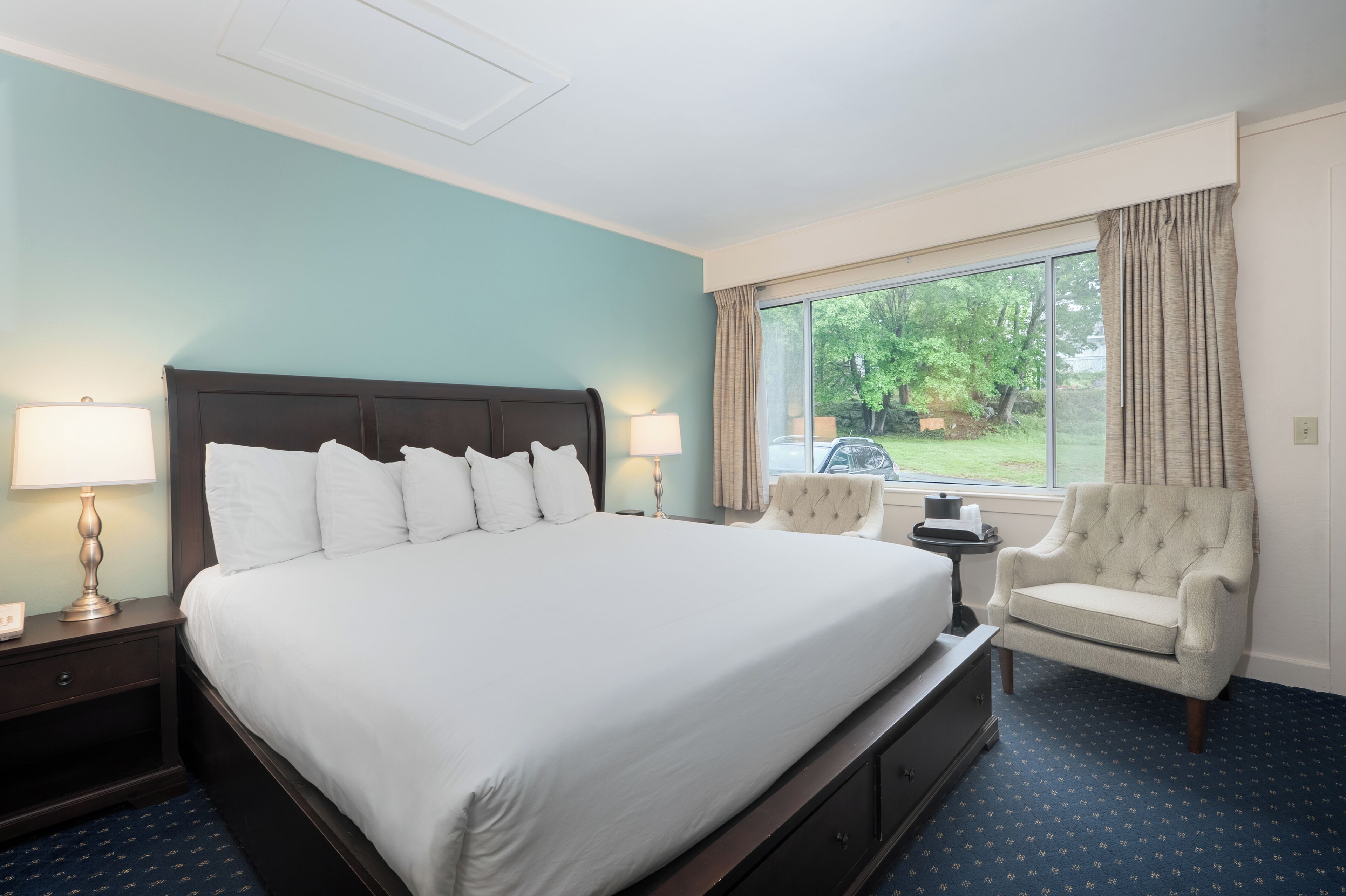 A hotel room with a large bed, two bedside lamps, a chair, a small table, and a window with curtains.