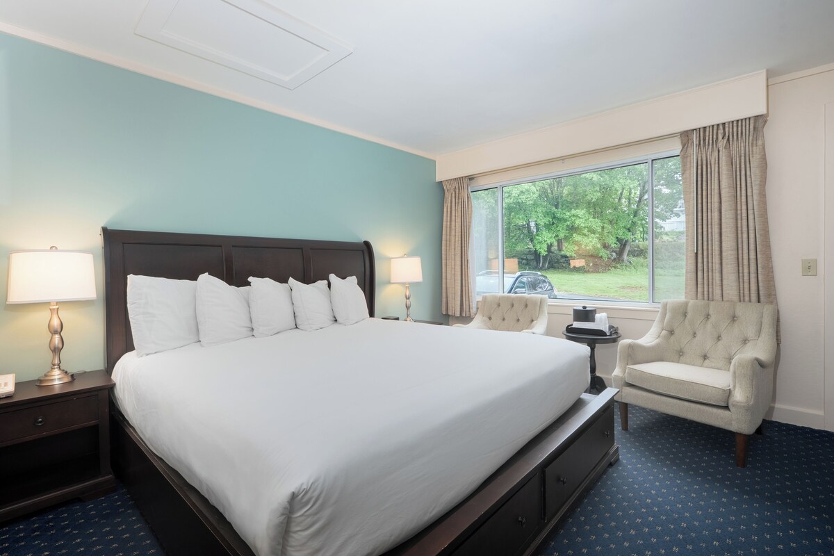 A hotel room with a large bed, two bedside lamps, a chair, a small table, and a window with curtains.