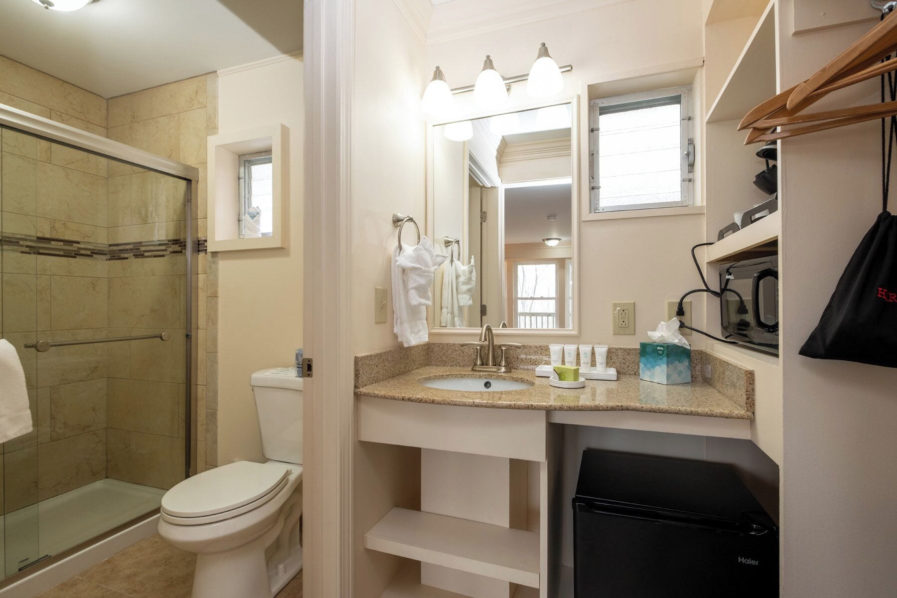 A bathroom with a glass shower enclosure, a toilet, a sink with a vanity, a mirror, and a black Haier refrigerator.