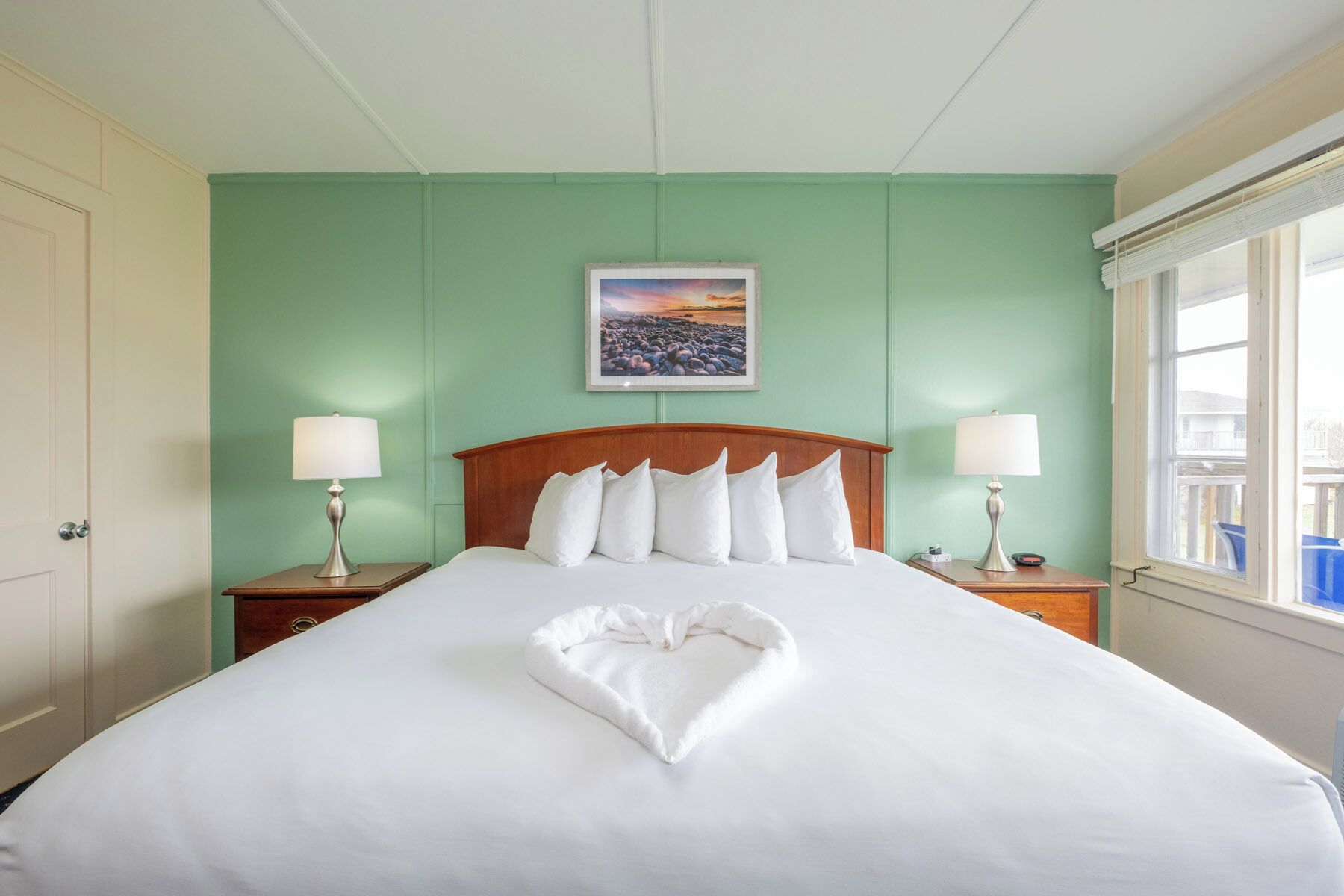 A neatly made bed with a heart-shaped towel, two bedside lamps, a framed picture on the wall, and a window with blinds.