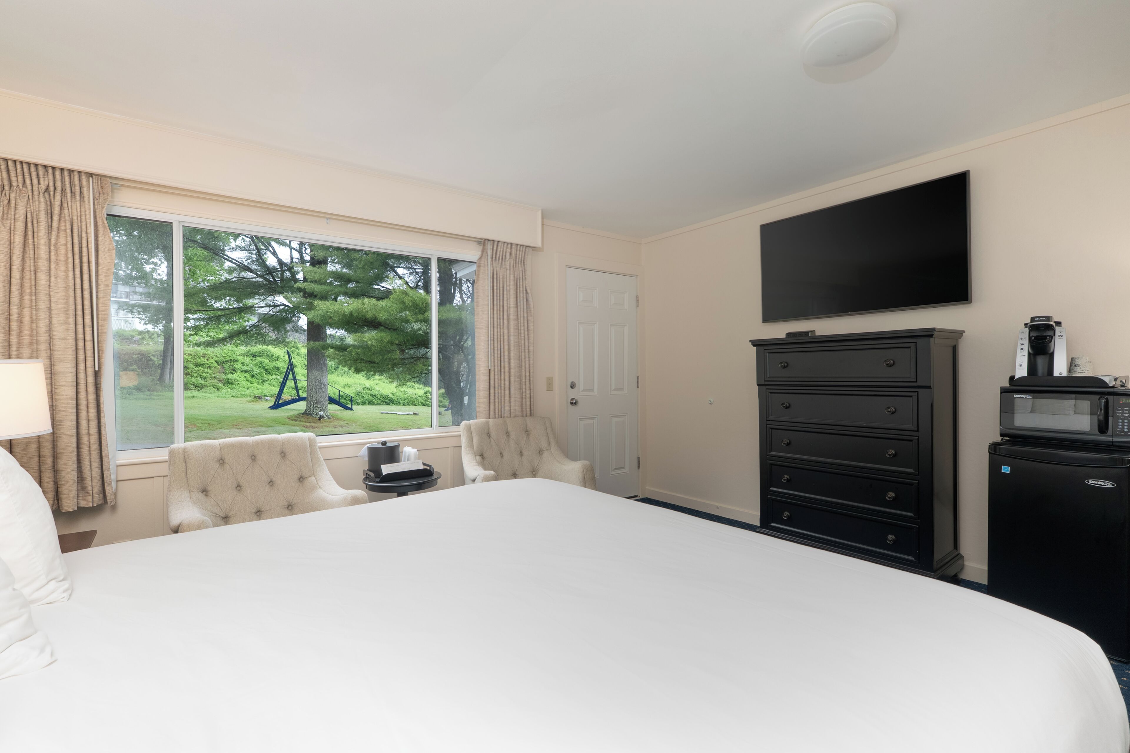 A hotel room with a large bed, a TV mounted on the wall, a dresser, and a window with curtains.