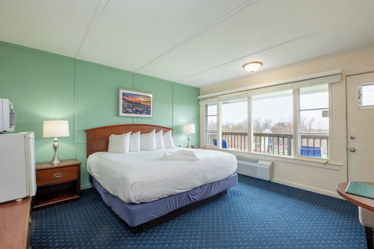 A hotel room with a bed, two bedside tables, a window with a view, and a painting on the wall.