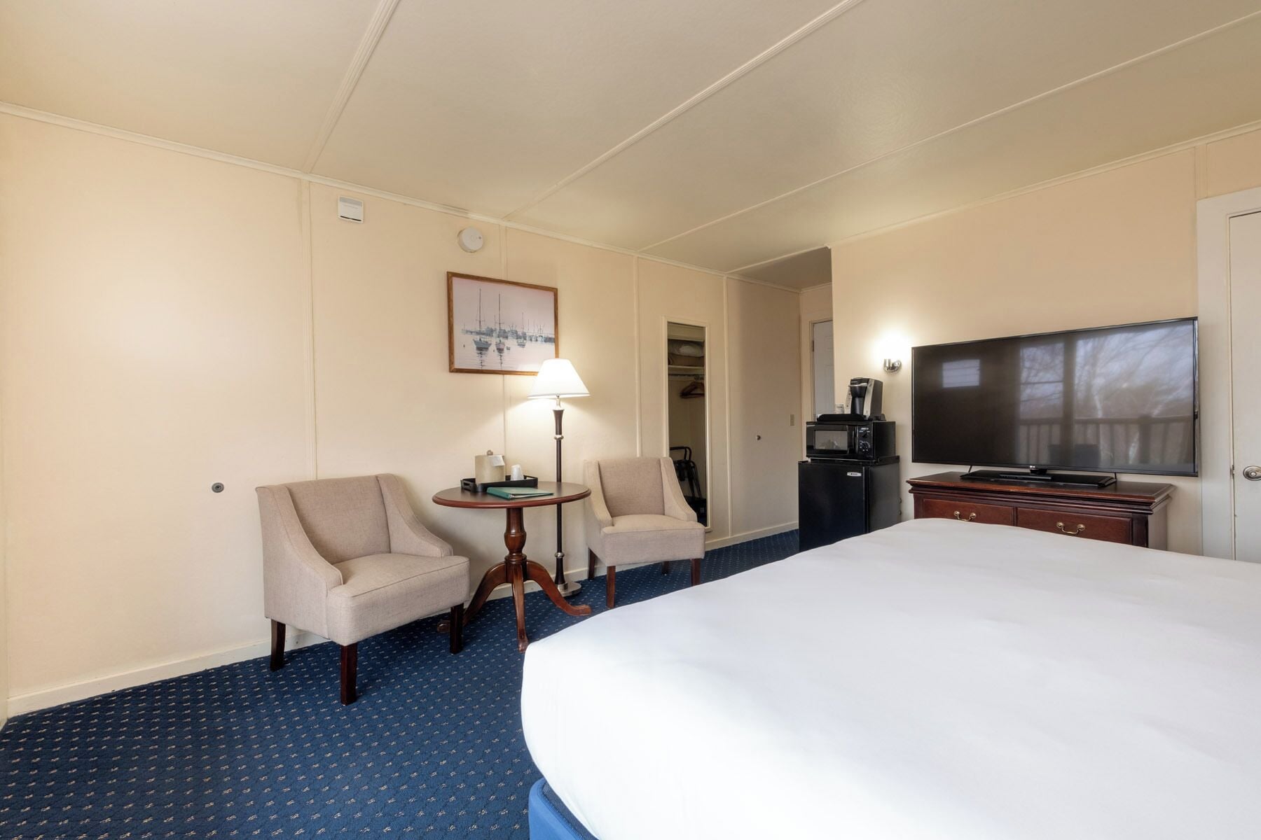 A hotel room with a large bed, two armchairs, a small table with a lamp, a television, and a closet.