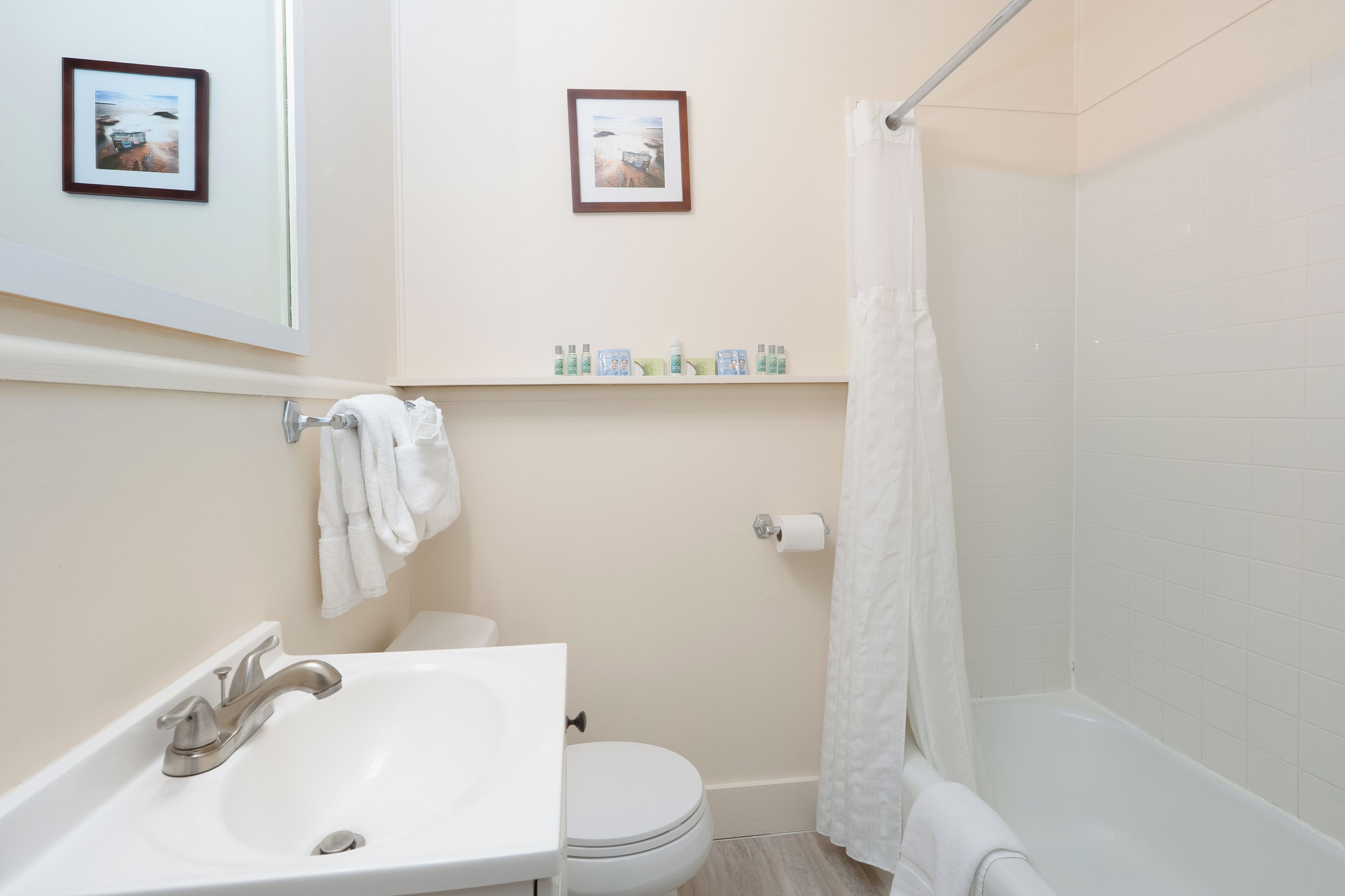 A bathroom with a white sink, toilet, and bathtub. There are two framed pictures on the wall, a towel rack with a towel, and a shelf with toiletries.