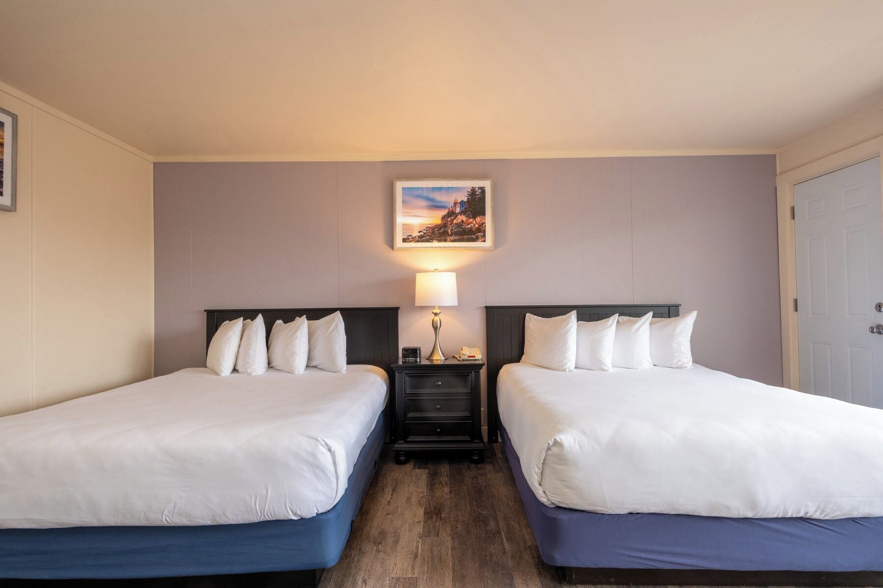 Una habitación de hotel con dos camas, una mesita de noche con lámpara, un cuadro en la pared y una puerta.