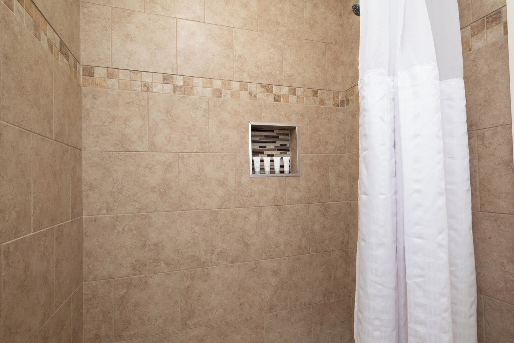 Un espace douche avec des carreaux de couleur beige, un rideau de douche blanc et une petite fenêtre où sont rangés les articles de toilette.