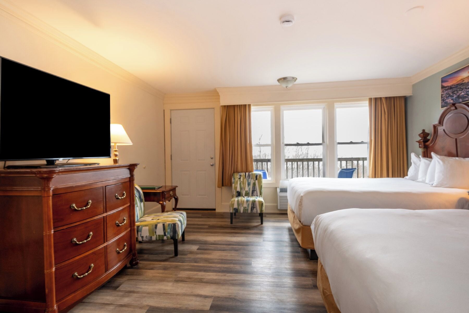 A hotel room with a TV, wooden dresser, two beds, a chair, and a balcony with a view.
