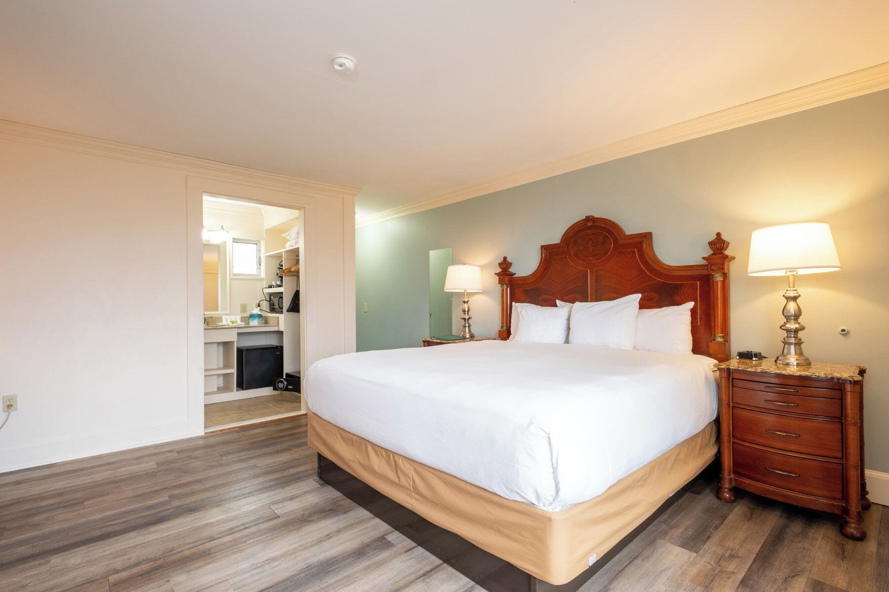 A hotel room with a large bed, wooden headboard, two bedside tables with lamps, a wardrobe, and a bathroom visible through an open door.
