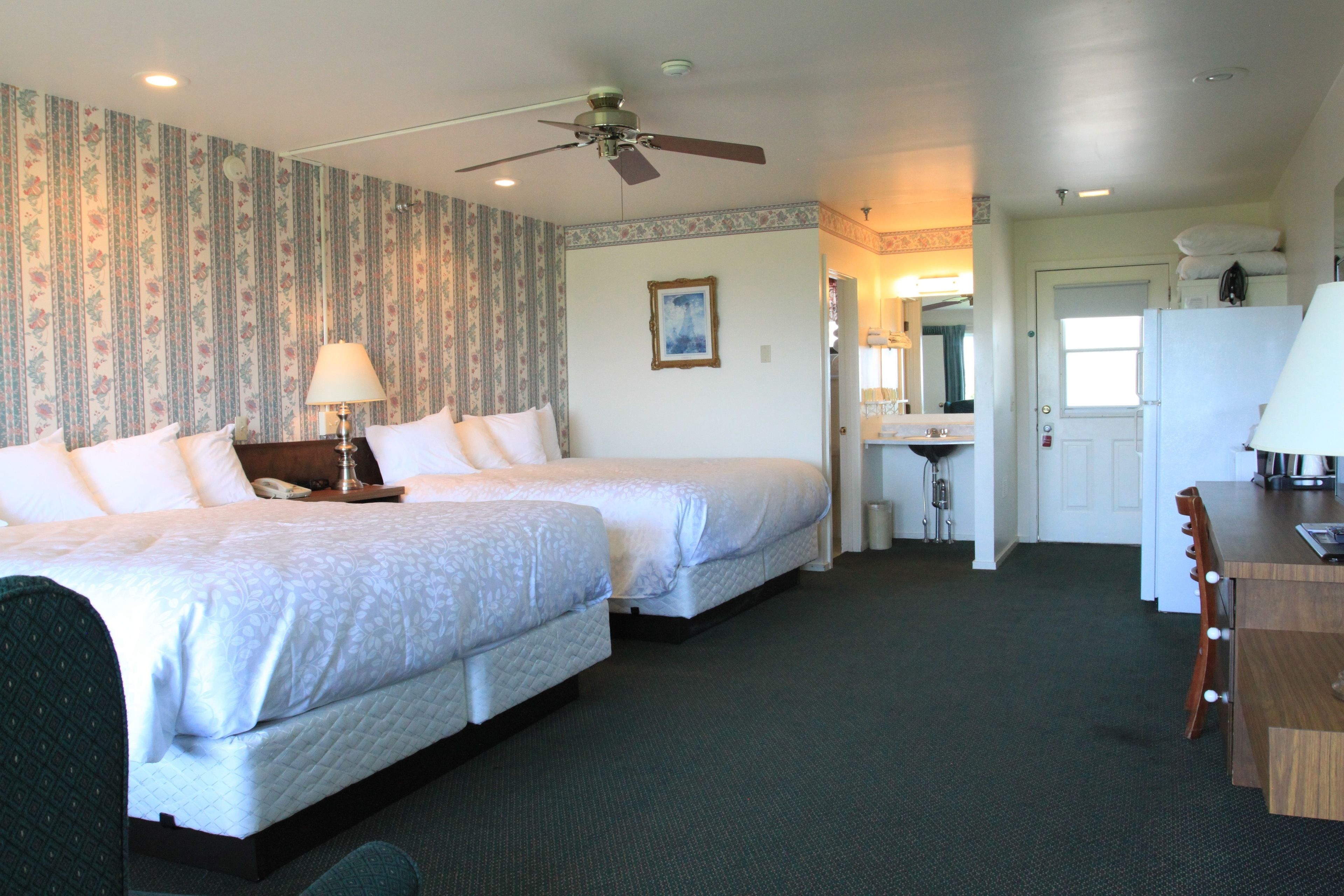 A hotel room with two beds, a desk, a chair, a ceiling fan, and a bathroom visible through an open door.