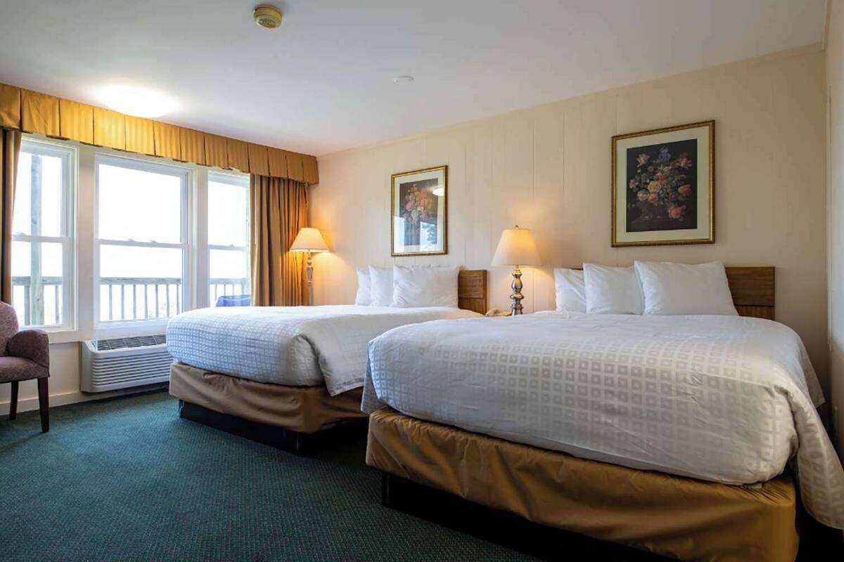 Habitación de hotel con dos camas, una silla, un ventanal con cortinas y un cuadro en la pared.