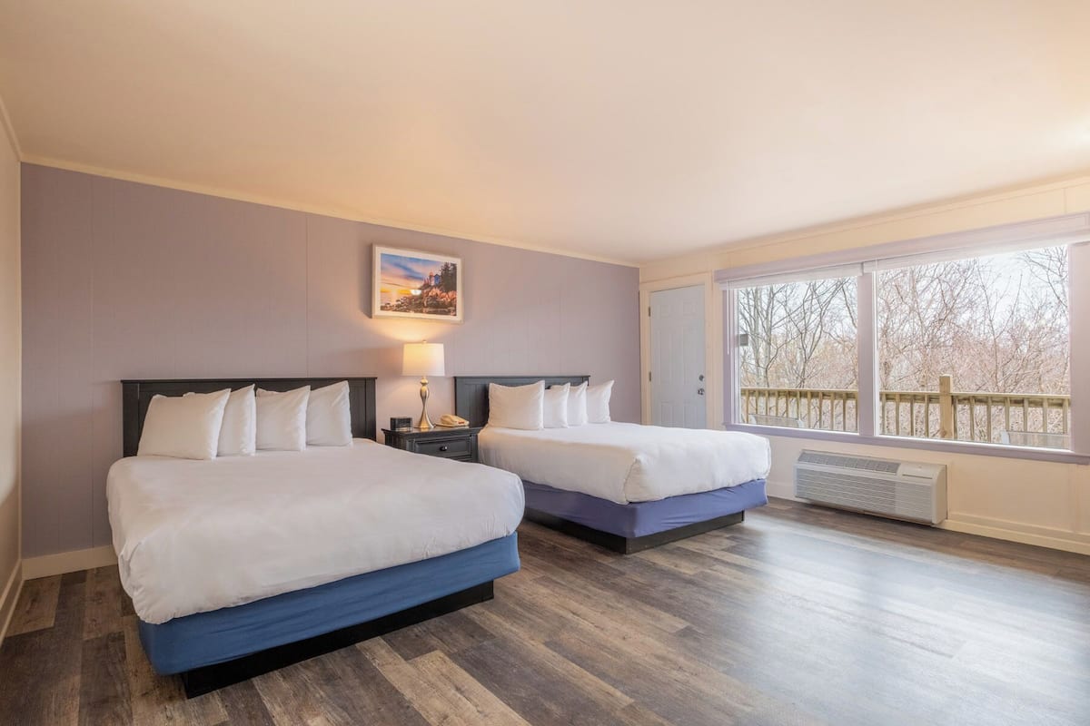 Habitación de hotel con dos camas, una mesita de noche, una lámpara, un cuadro y un ventanal con vistas a árboles.