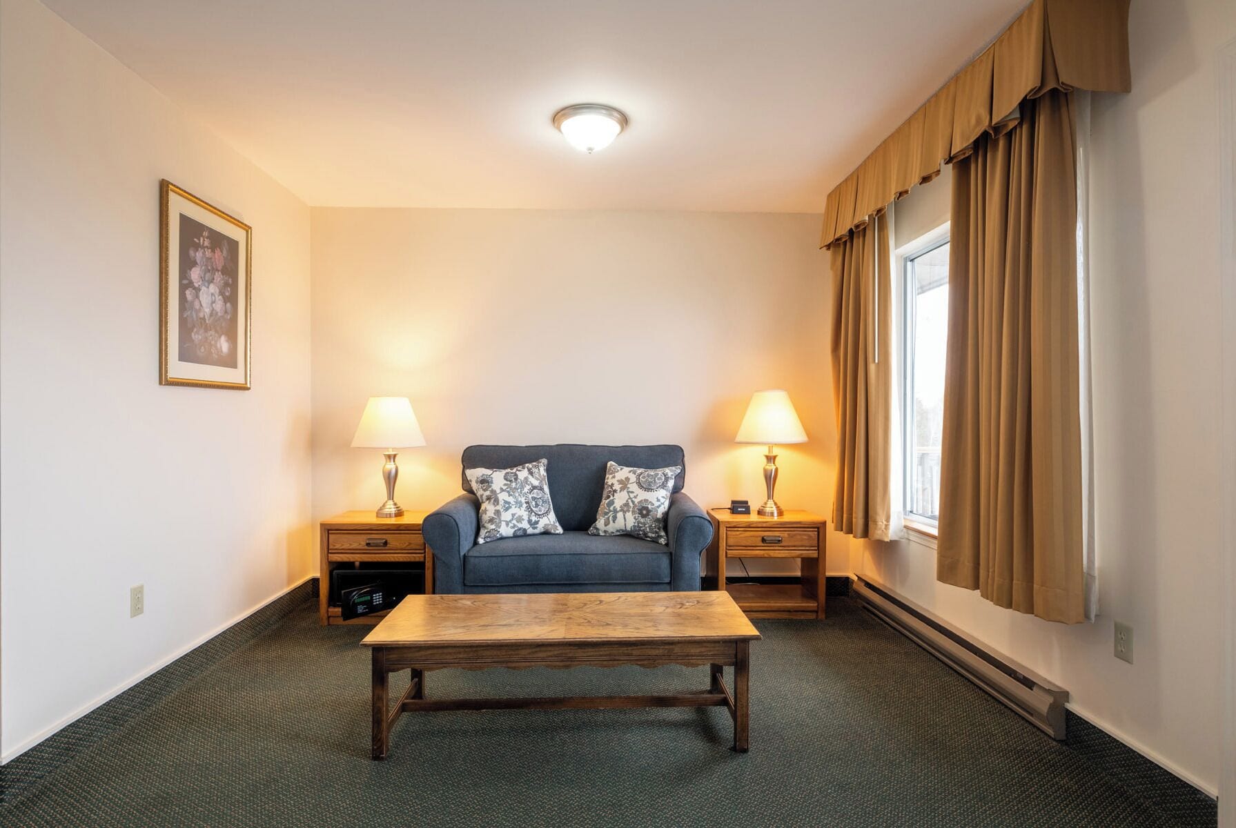 Une chambre d’hôtel avec un canapé bleu, deux tables de chevet avec des lampes, une table basse en bois et un tableau encadré au mur.