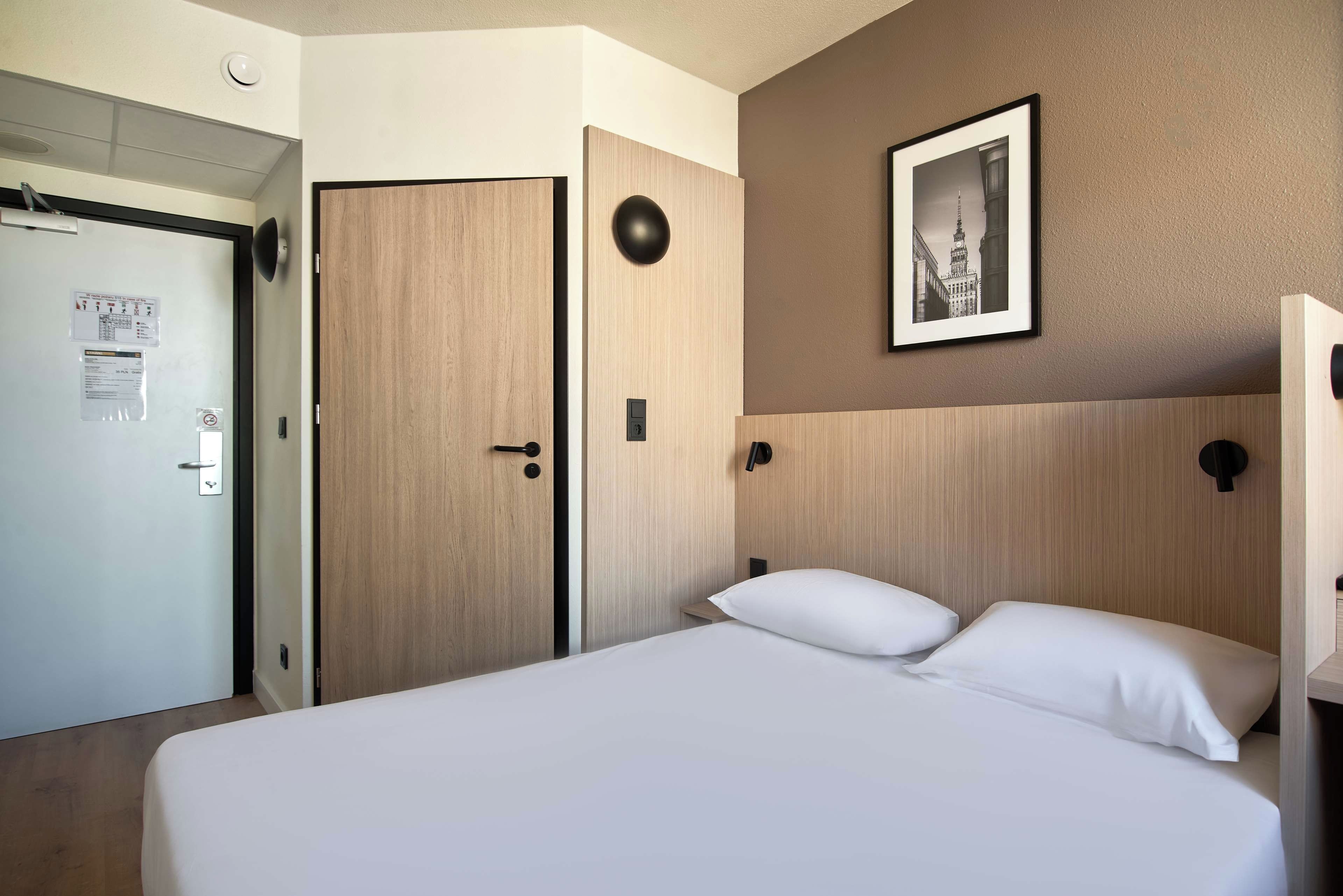 A hotel room with a bed, a wardrobe, a door, a wall-mounted light, and a framed picture.