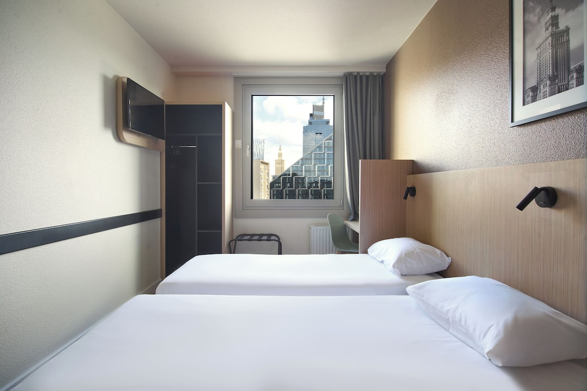 A hotel room with two beds, a window with a city view, a desk, and a TV mounted on the wall.