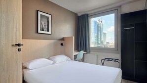 A hotel room with a bed, a desk, a chair, a framed picture on the wall, and a window with a city view. - Premiere Classe Warszawa Centrum (Warsaw)