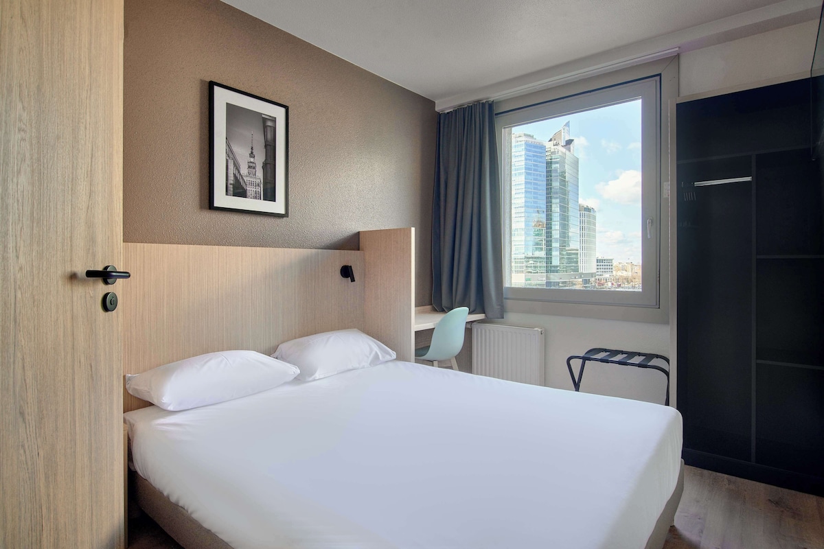 A hotel room with a bed, a desk, a chair, a framed picture on the wall, and a window with a city view.