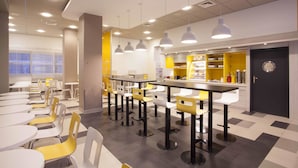 A modern cafeteria with round tables and chairs, a kitchen area, and a black door with a circular emblem. - Premiere Classe Warszawa Centrum (Warsaw)