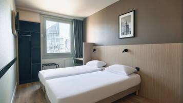 Une chambre d’hôtel avec deux lits, une fenêtre donnant sur des immeubles, une porte et un tableau encadré au mur.