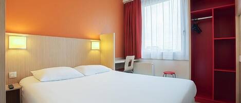 A hotel room with a large bed, a desk, a chair, a wardrobe, and a window with curtains.