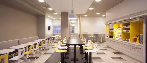 Una cafetería moderna con mesas redondas y sillas amarillas, una zona central de servicio y un mostrador de recepción.