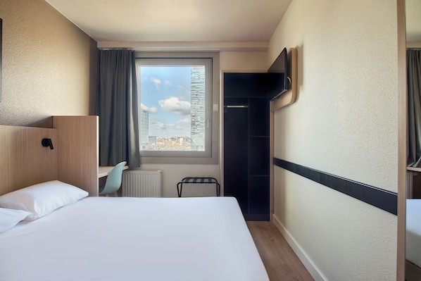 Camera d'albergo con un letto, una finestra che dà su edifici, un termosifone, una sedia e una TV fissata al muro.