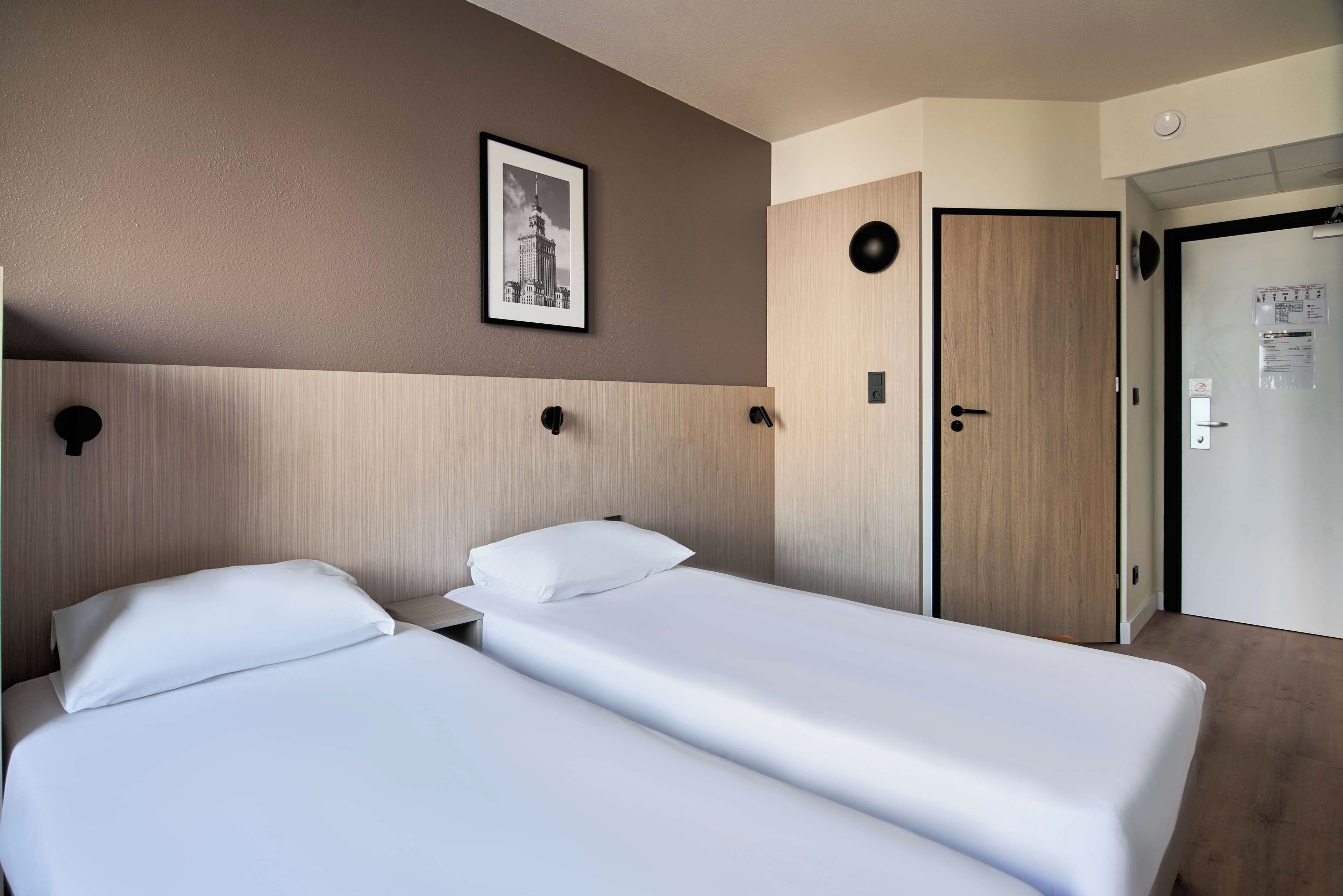 Une chambre d’hôtel avec deux lits simples, une tête de lit en bois, un tableau encadré au mur et une porte dotée d’un interrupteur et d’une petite pancarte.