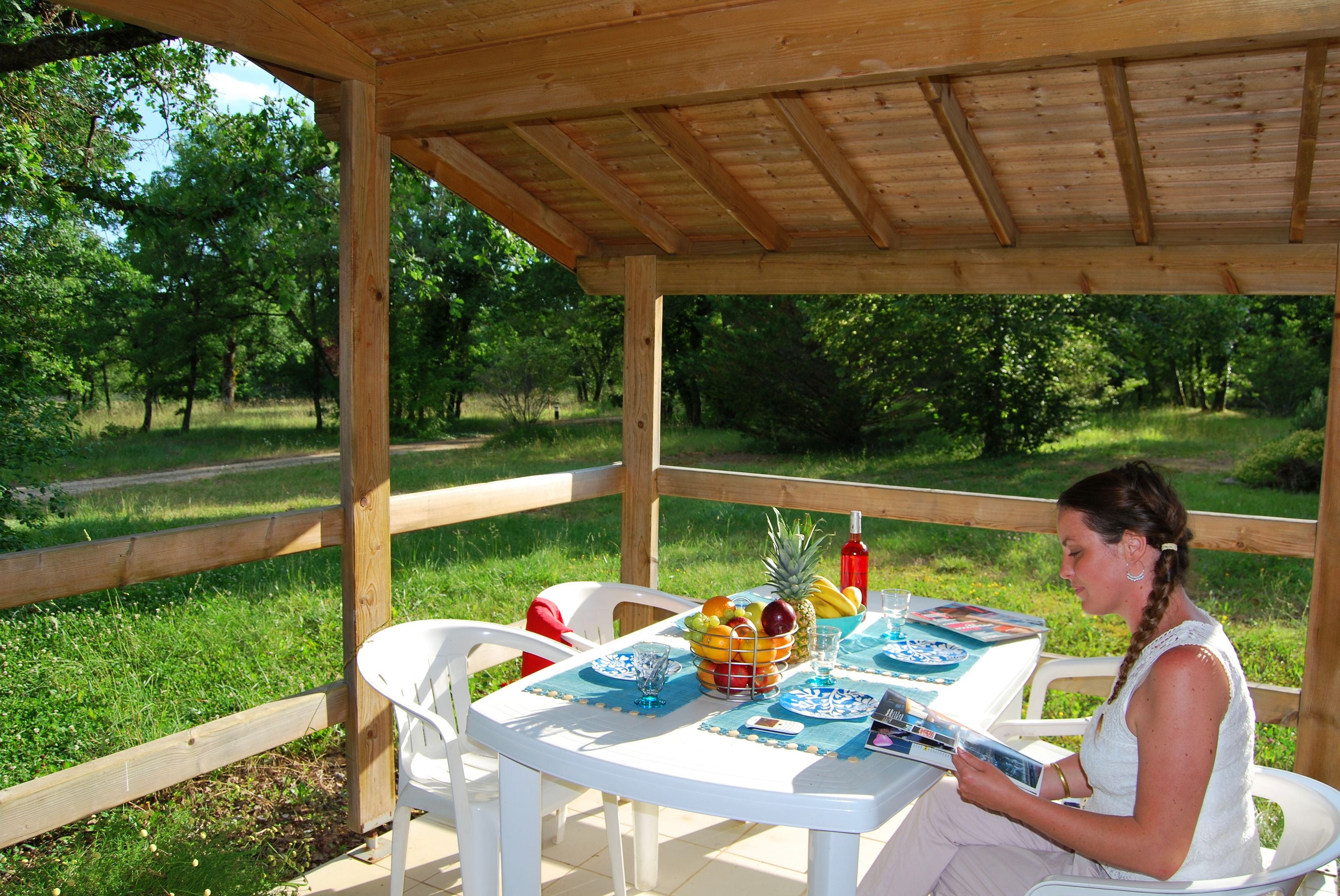Ein überdachter Essbereich im Freien mit einer Holz-Pergola, weißen Stühlen und einem Tisch, der mit Früchten, einer Flasche und einer Speisekarte gedeckt ist.