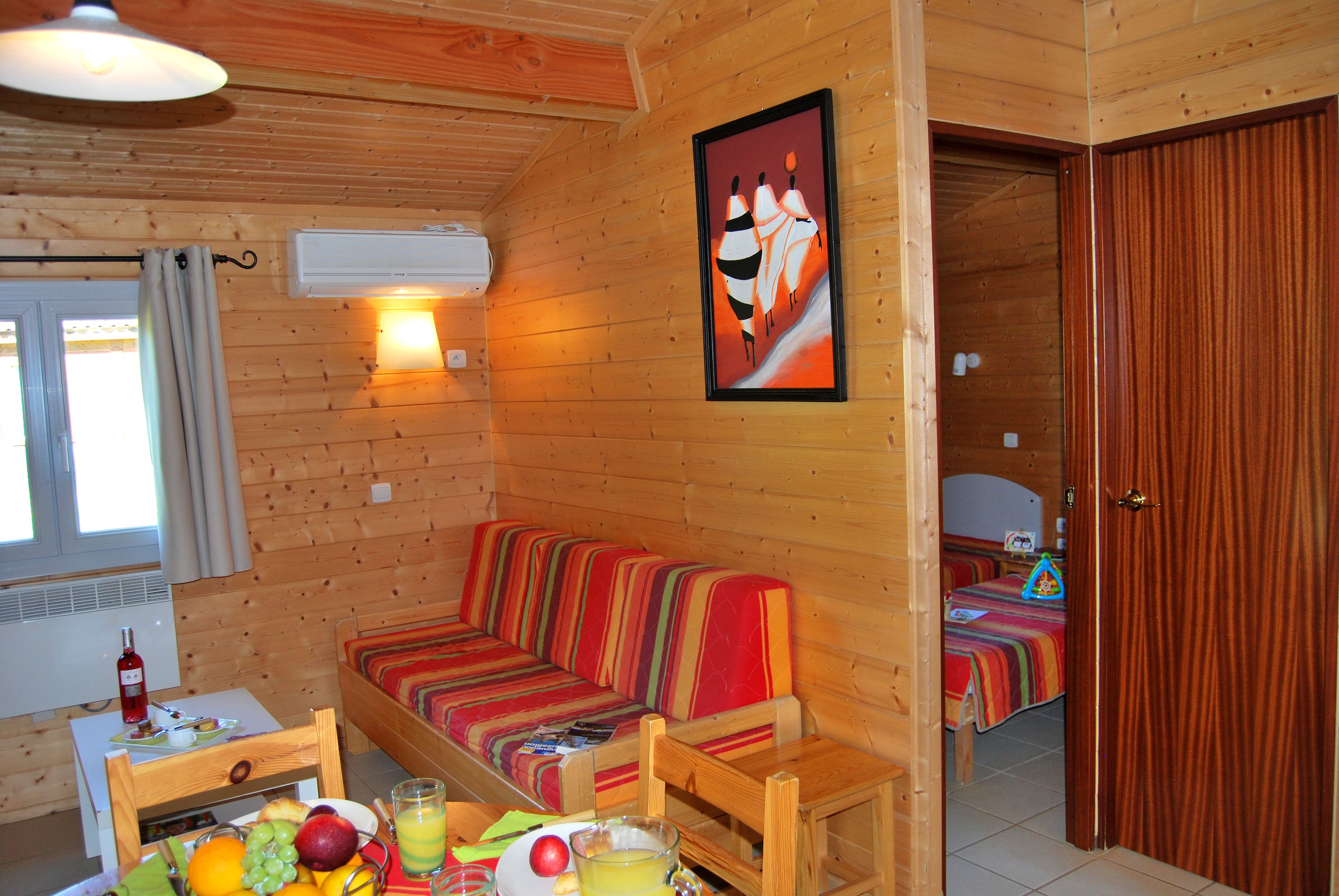 A cozy wooden cabin with a wooden sofa, a dining table with fruits and a glass of juice, and a bed with a striped blanket.