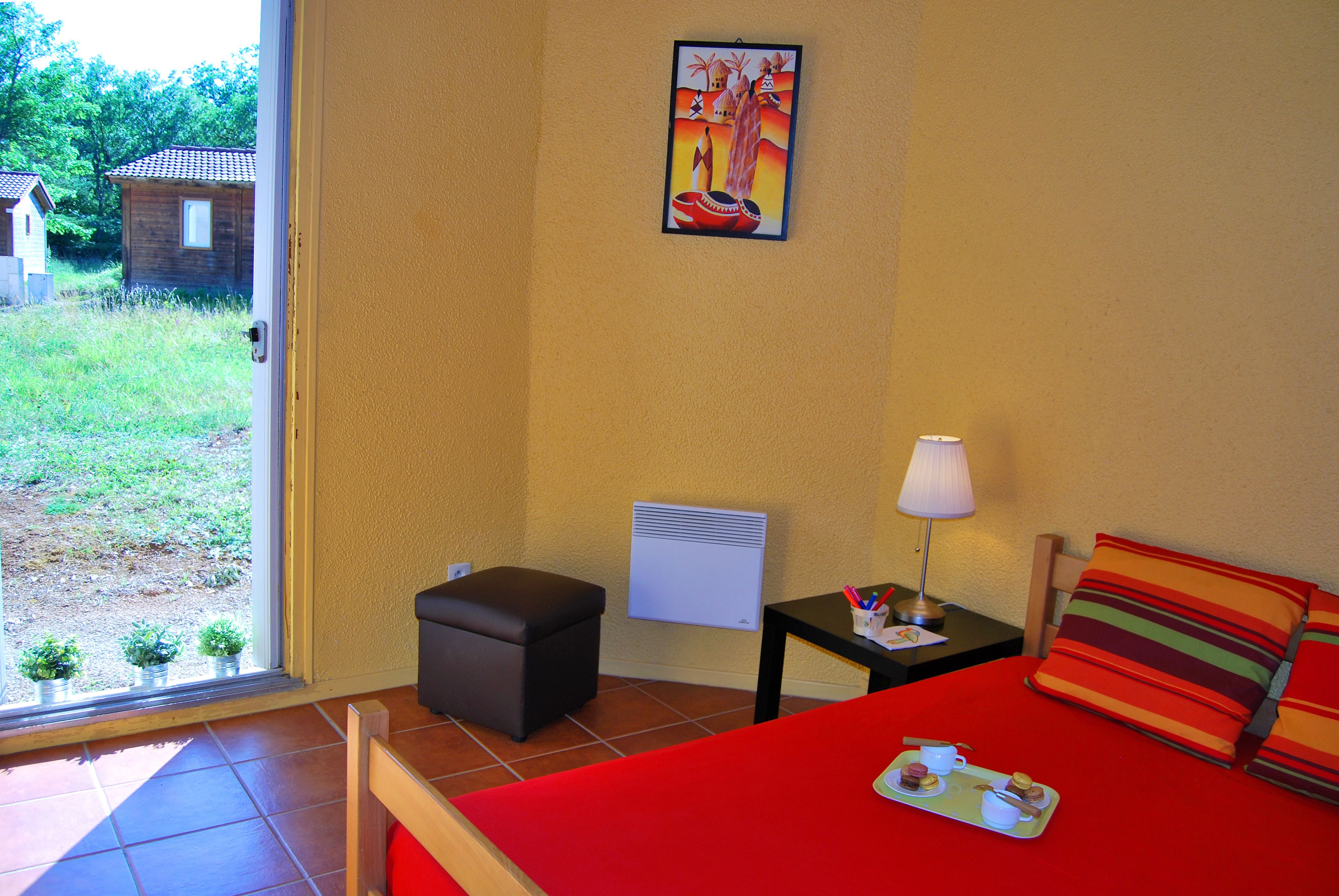 Une pièce avec un lit rouge, une petite table avec une lampe, une chaise, une fenêtre donnant sur l’extérieur et un tableau accroché au mur.