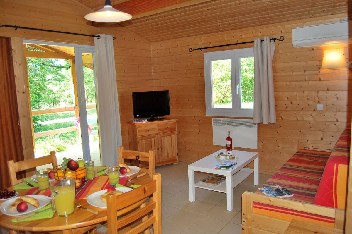 A wooden cabin with a dining table, chairs, a sofa, a television, and a window overlooking greenery.