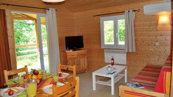 A wooden cabin with a dining table, chairs, a sofa, a television, and a window overlooking greenery.