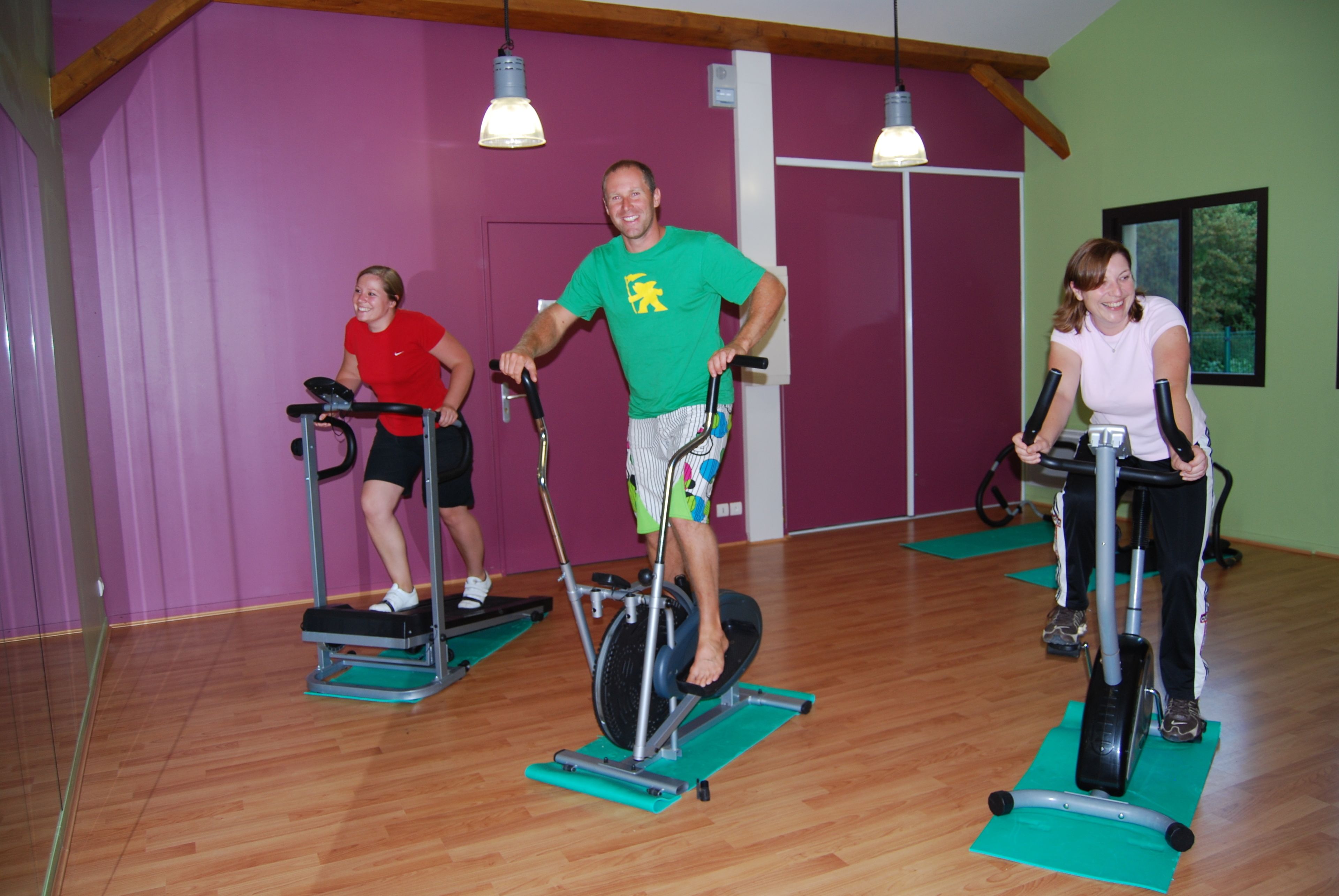 Three people using stationary bikes in a room with wooden floors and purple walls.