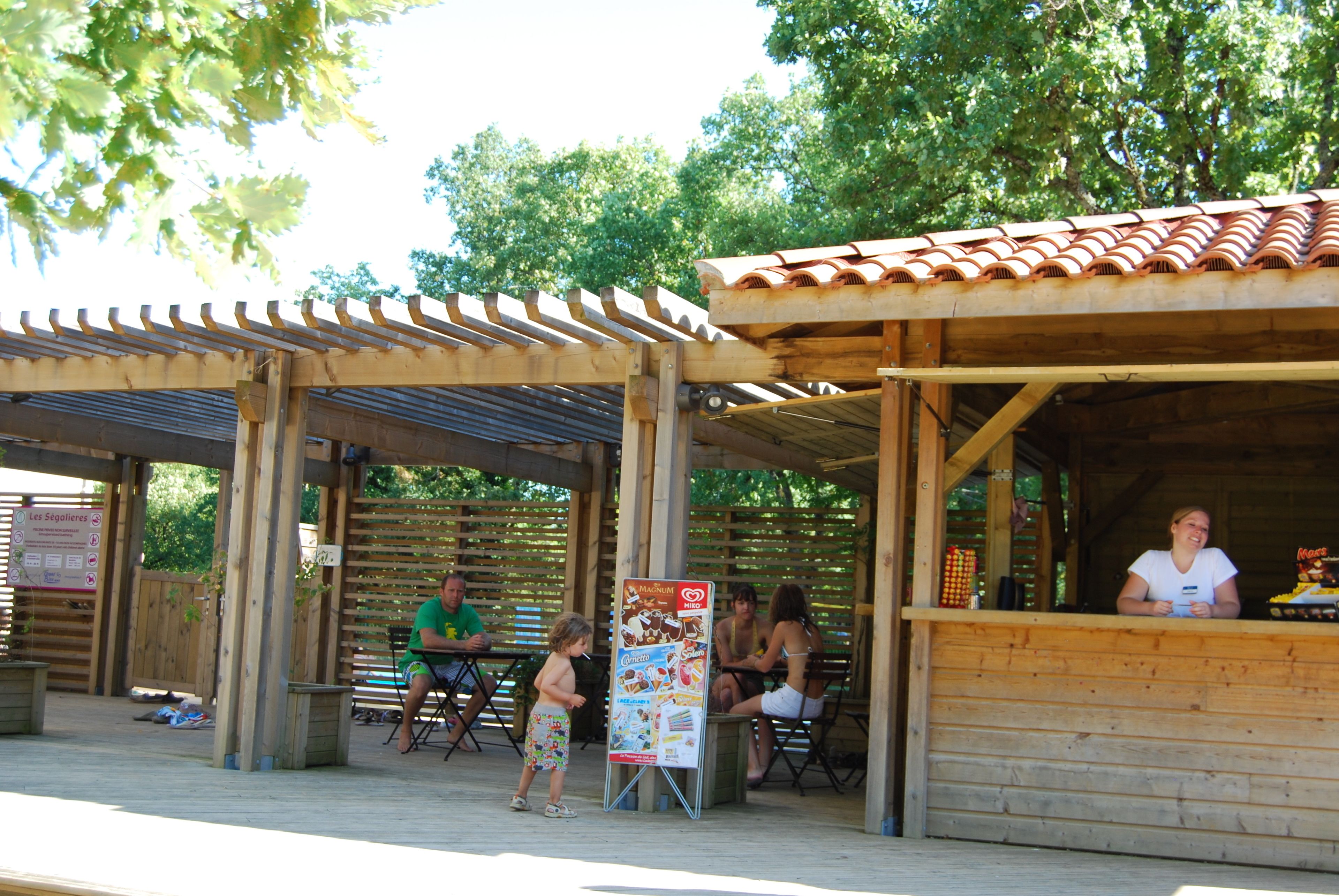Un espace extérieur aménagé avec des tables et des chaises en bois, une partie couverte avec un toit en tuiles et un panneau indicateur.